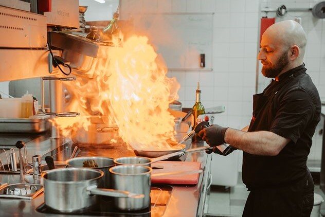 A chef is standing in the kitchen of the Ganymed Brasserie, flambéing a dish.